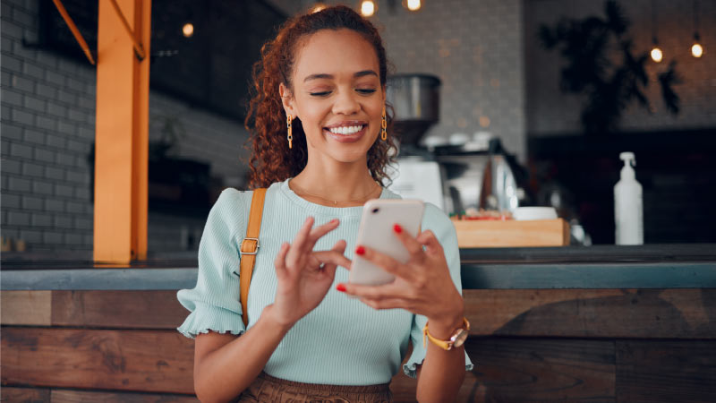 A woman standing indoors, smiling while using a smartphone. She wears a light blue top and has curly hair. In the background, there is a counter and some blurred lights.